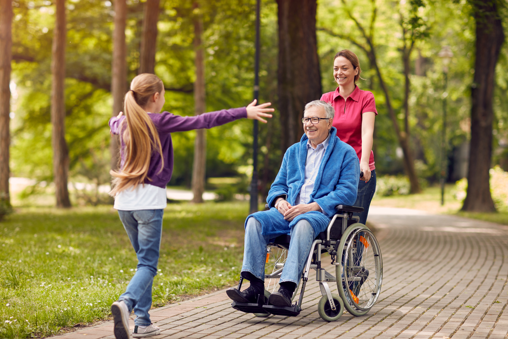 A young girl greets her grandfather in his wheelchair, pushed by her mother